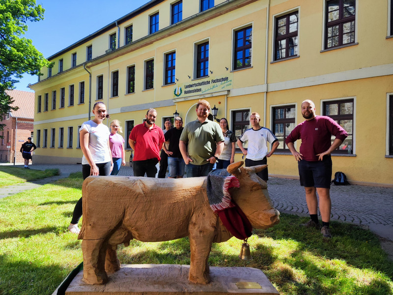 Zu sehen sind Schüler, sie stehen vor einer symbolischen Holzkuh vor dem Gebäude der Fachschule Haldensleben. 