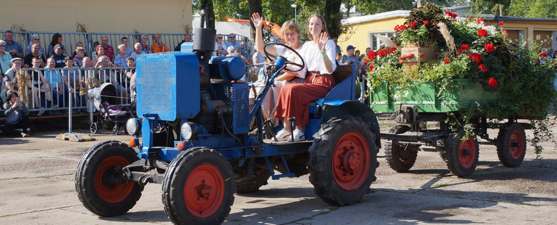 Zu sehen ist ein Traktor, bunt geschmückt mit Blumen. Es ist ein Oldtimer und zwei Frauen sitzen darauf. Sie winken dem Publikum am Rand stehend zu. 