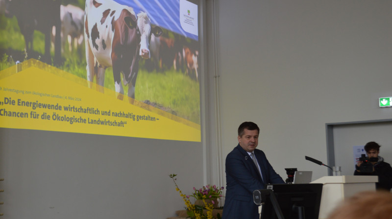 Das Bild zeigt den Landwirtschaftsminister des Landes Sachsen-Anhalt Sven Schulze während seiner Rede zur Jahrestagung Ökolandbau. Im Hintergrund zu sehen ist ein großes Motto-Bild mit einer Kuh. 